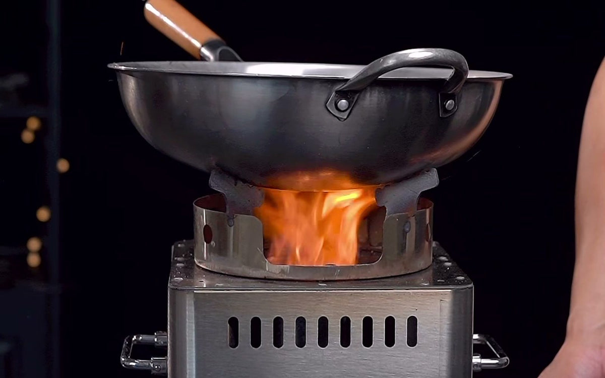 Hillrein Zhengtu wood stove for wok cooking with a dark two-handled wok and wooden-handled utensil above an orange flame